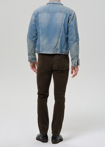 A man viewed from the back wearing a light blue denim jacket, dark brown slim-fit pants, and black leather shoes, standing against a grey background.