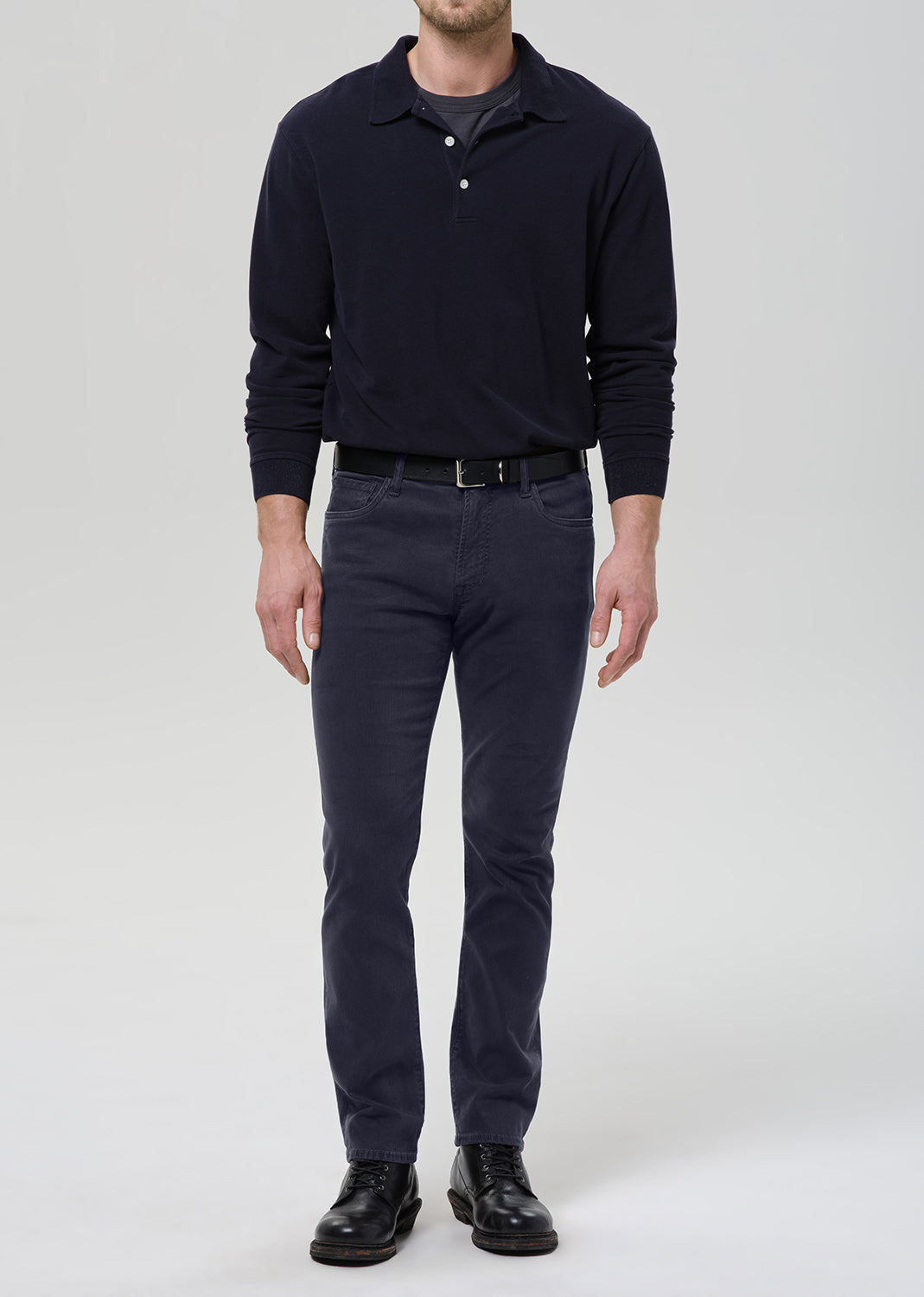 A man wearing a dark navy long-sleeve polo shirt, dark gray slim-fit trousers, a black belt, and black leather shoes, standing against a grey background.
