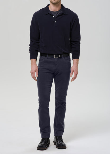 A man wearing a dark navy long-sleeve polo shirt, dark gray slim-fit trousers, a black belt, and black leather shoes, standing against a grey background.