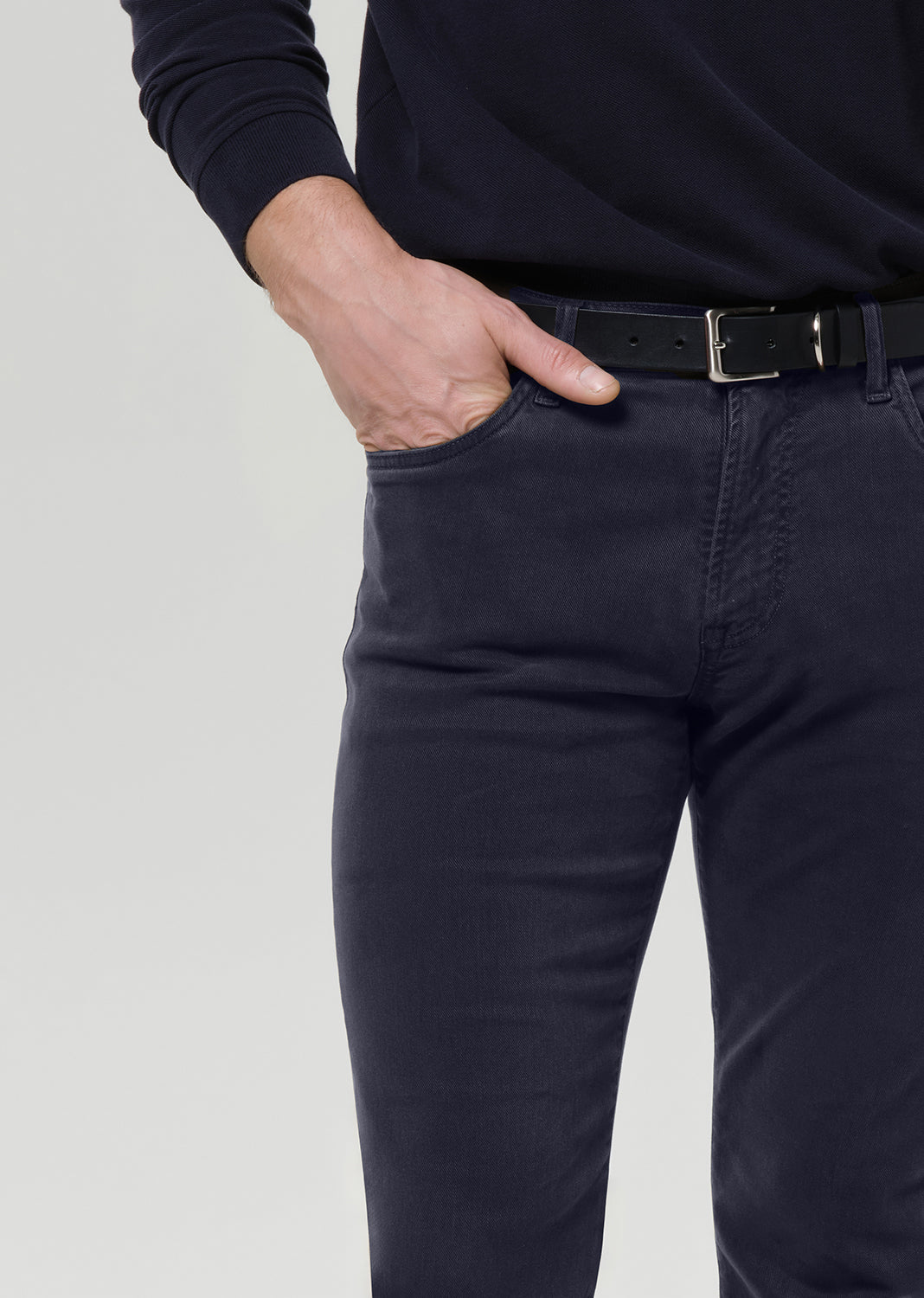 Close-up of a person wearing dark navy slim-fit jeans with a black belt, paired with a black long-sleeve top, against a grey background.