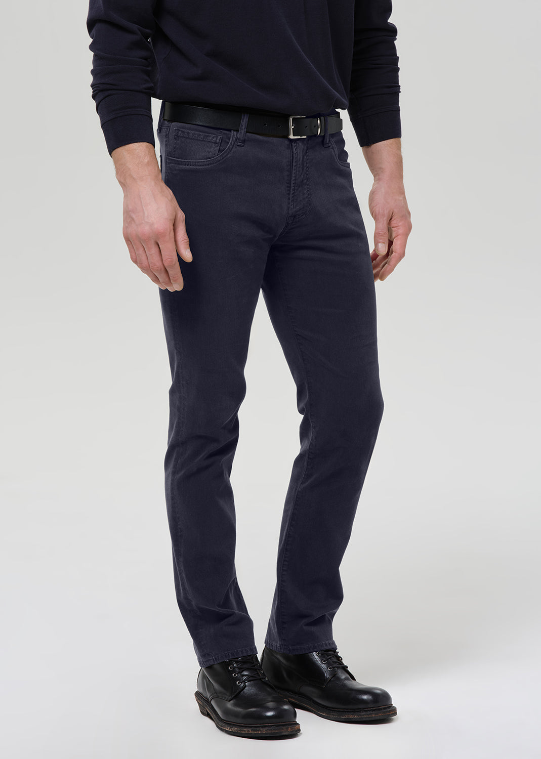 A man wearing dark navy slim-fit jeans, a black belt, a dark navy long-sleeve shirt, and black leather shoes, standing against a grey background.