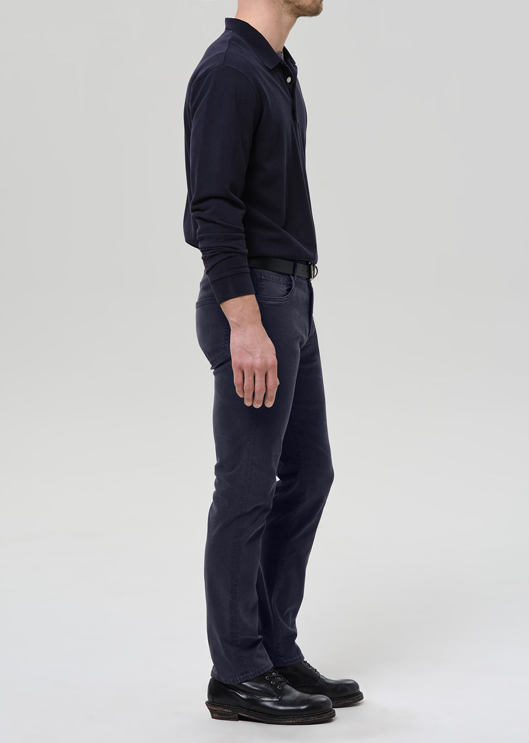 A man shown in profile wearing a dark navy long-sleeve polo shirt, matching navy trousers, a black belt, and black leather shoes against a grey background.