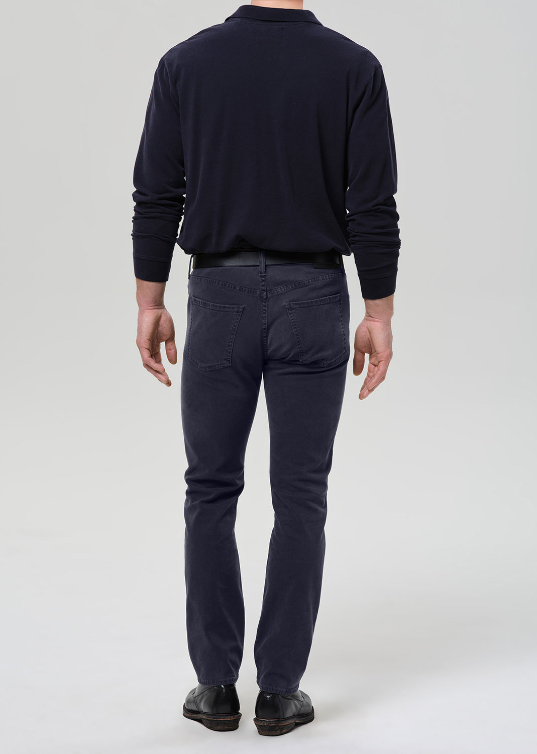 A man viewed from the back wearing a dark navy long-sleeve shirt, dark navy pants, and black shoes, standing against a grey background.