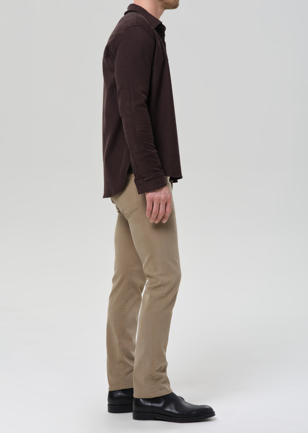 A man shown in profile wearing a dark brown long-sleeve button-up shirt, beige trousers, and black leather shoes, photographed against a grey background.