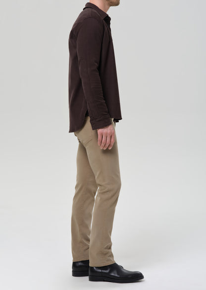 A man shown in profile wearing a dark brown long-sleeve button-up shirt, beige trousers, and black leather shoes, photographed against a grey background.