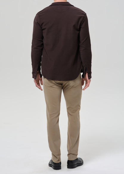 A man viewed from the back wearing a dark brown long-sleeve button-up shirt, beige trousers, and black shoes, standing against a greybackground.