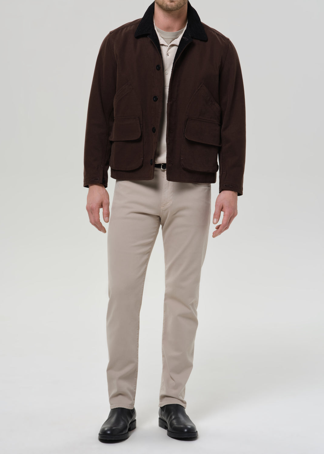A man modeling a dark brown corduroy jacket with a black corduroy collar, worn over a beige t-shirt and paired with beige trousers and black leather shoes, against a grey background.