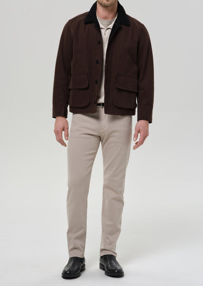 A man modeling a dark brown corduroy jacket with a black corduroy collar, worn over a beige t-shirt and paired with beige trousers and black leather shoes, against a grey background.