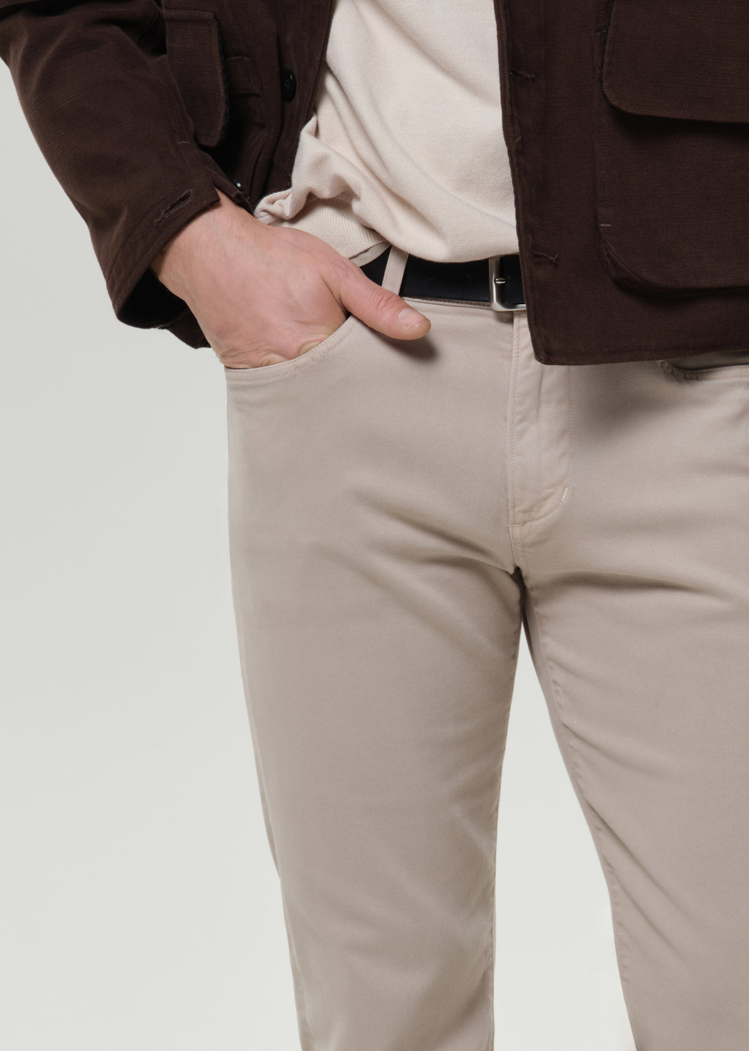 A close-up of a person wearing light beige chinos with a black belt, a cream shirt, and a brown jacket, with one hand in the pocket.