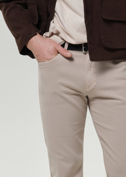 A close-up of a person wearing light beige chinos with a black belt, a cream shirt, and a brown jacket, with one hand in the pocket.
