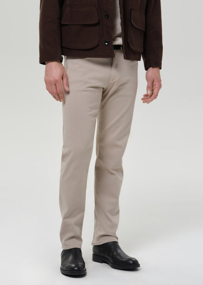 A model from the chest down wearing beige slim-fit trousers, a dark brown jacket, a light-colored shirt, and black leather boots, photographed against a grey background.
