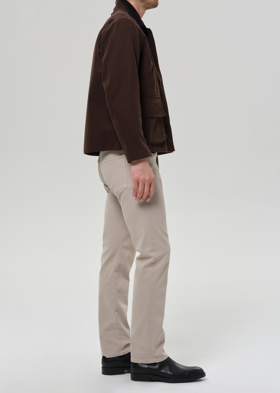 A man shown from the side wearing a dark brown corduroy jacket, beige trousers, and black shoes, standing against a grey background.
