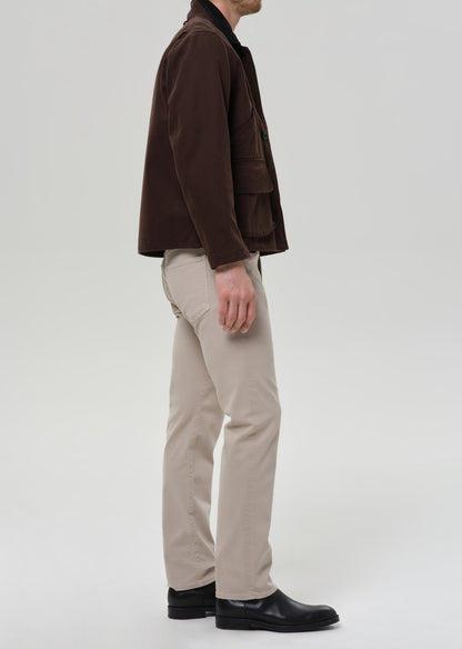 A man shown from the side wearing a dark brown corduroy jacket, beige trousers, and black shoes, standing against a grey background.