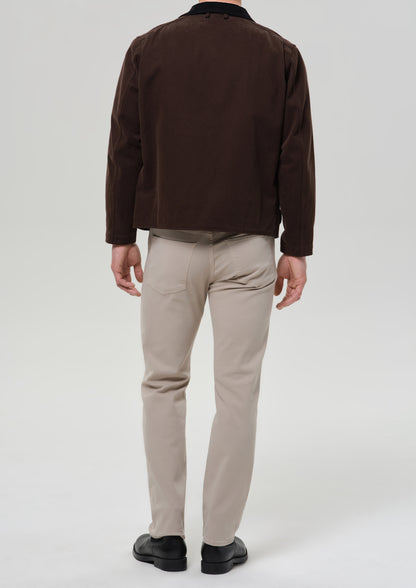 A man viewed from the back wearing a dark brown jacket, beige trousers, and black shoes, standing against a grey background.