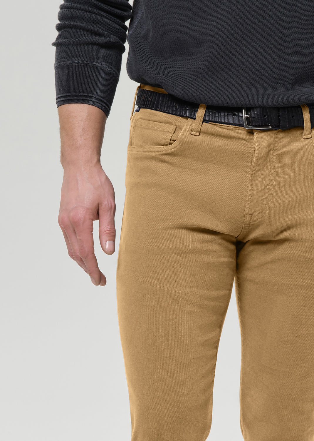 A close-up of a person wearing tan chinos with a black textured belt and a dark charcoal ribbed sweater, cropped from the chest down against a light grey background.