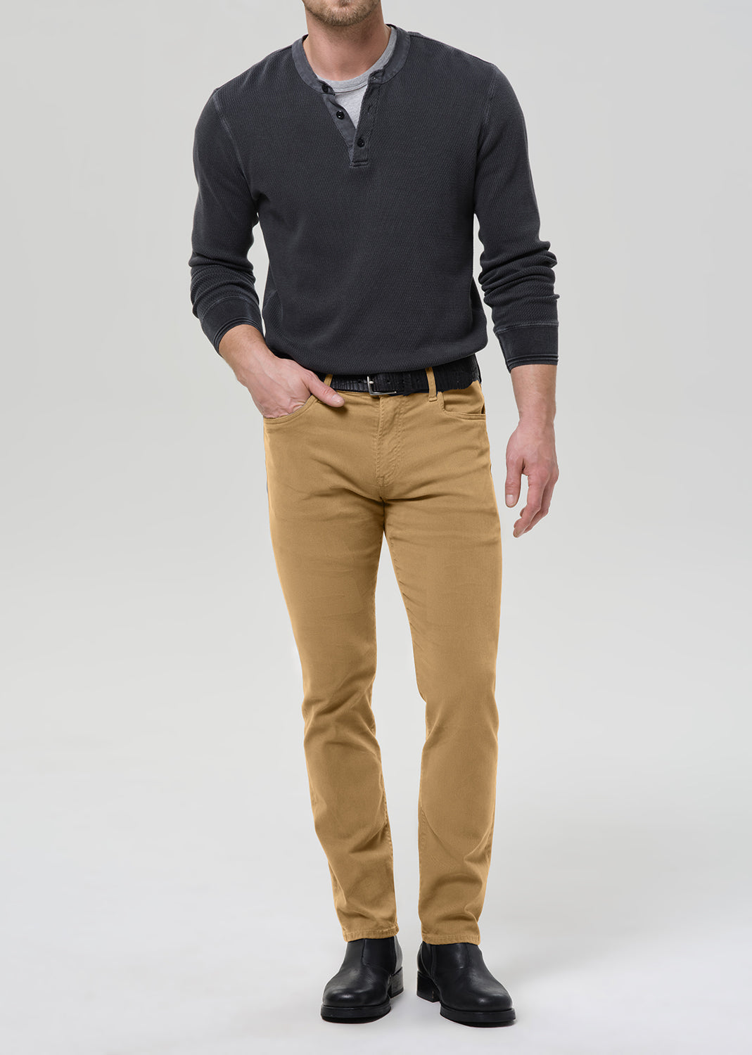 A man modeling slim-fit tan chinos with a black belt, a dark charcoal henley shirt, and black boots, photographed against a grey background.
