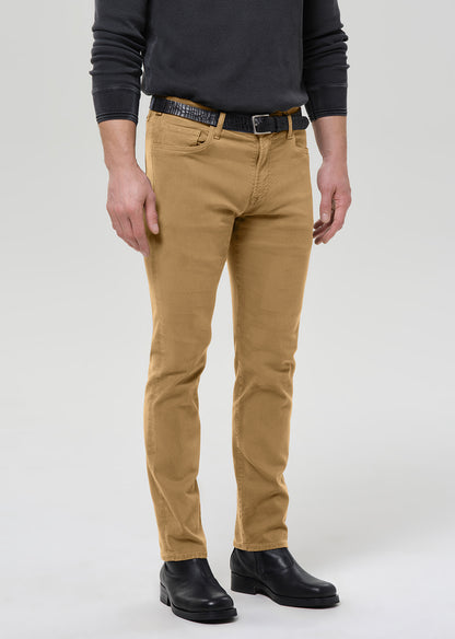 A man wearing slim-fit tan chinos with a black belt, black long-sleeve shirt, and black leather boots, standing against a grey background.
