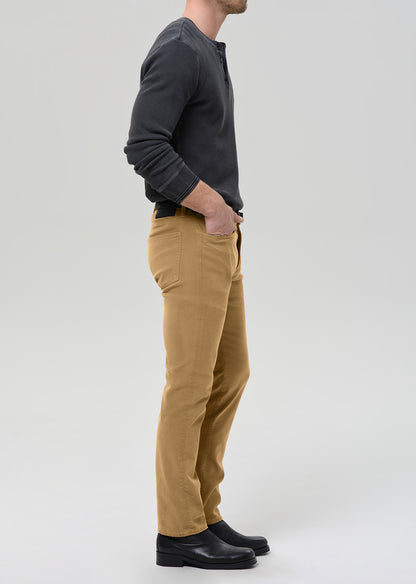 A man shown in profile wearing tan chinos, a dark charcoal henley shirt, and black leather shoes, standing against a grey background.