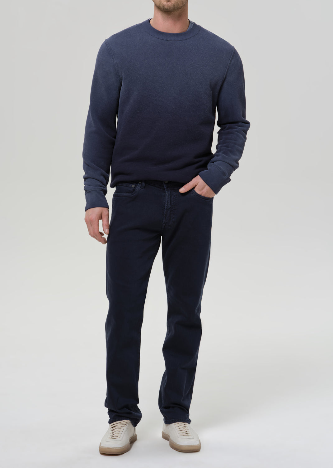 A man wearing a navy blue crewneck sweater and matching navy blue jeans, paired with beige sneakers, standing against a grey background.