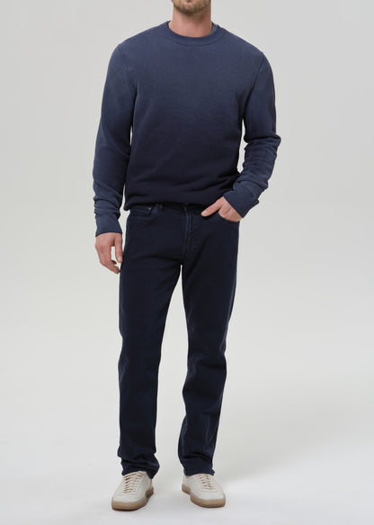 A man wearing a navy blue crewneck sweater and matching navy blue jeans, paired with beige sneakers, standing against a grey background.