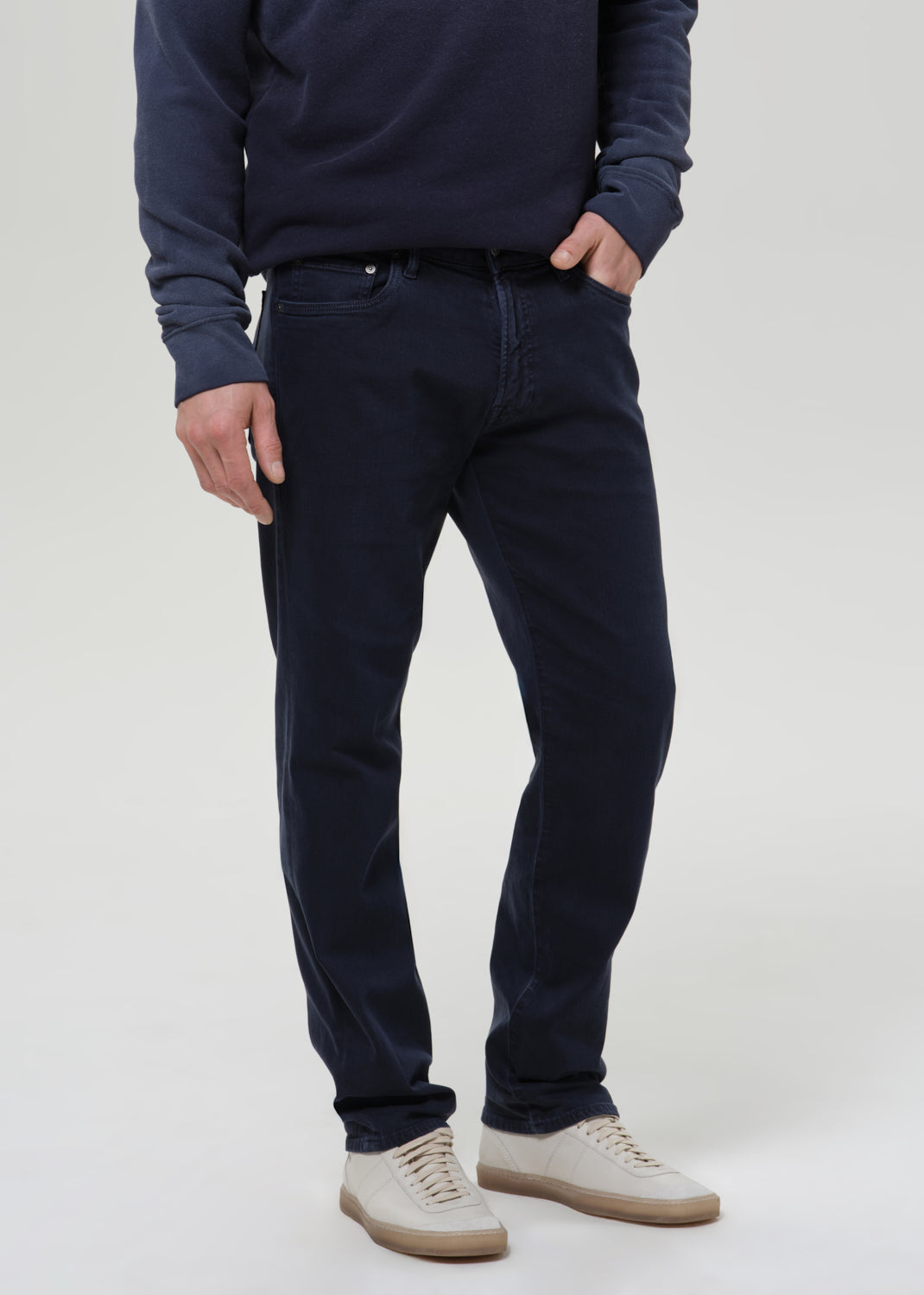 A man wearing dark navy blue slim-fit jeans, a matching navy sweatshirt, and off-white sneakers, standing against a grey background.