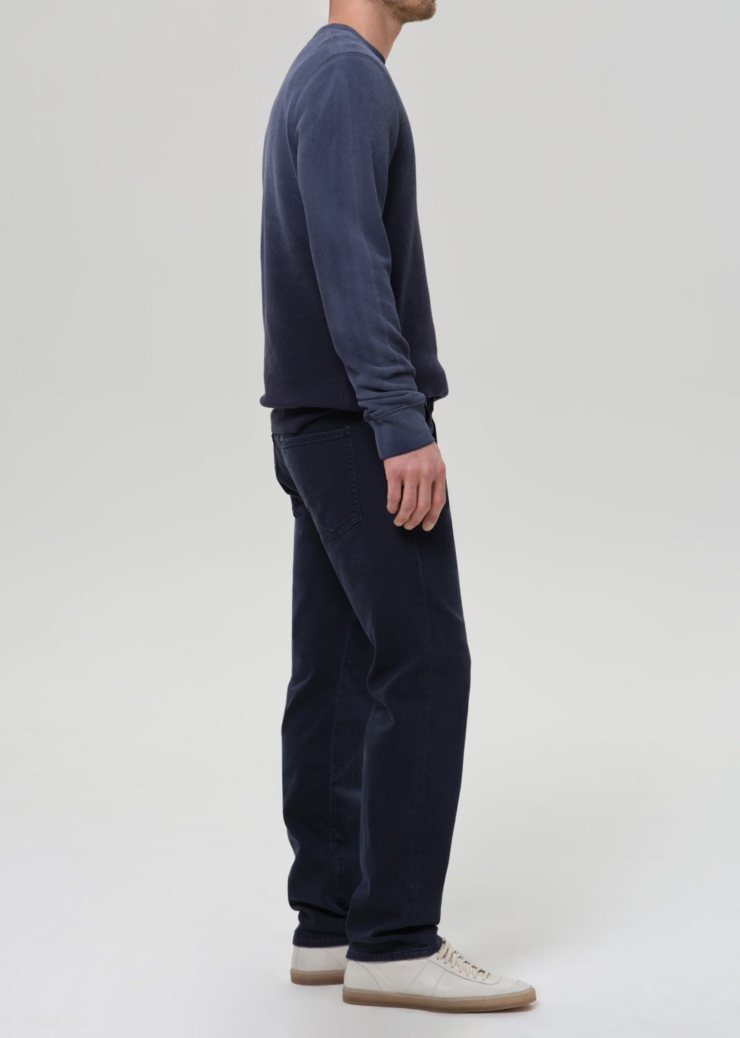 A man shown from the side wearing a dark navy long-sleeve crewneck sweatshirt, matching dark navy pants, and off-white sneakers, photographed against a grey background.