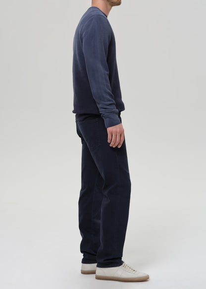 A man shown from the side wearing a dark navy long-sleeve crewneck sweatshirt, matching dark navy pants, and off-white sneakers, photographed against a grey background.