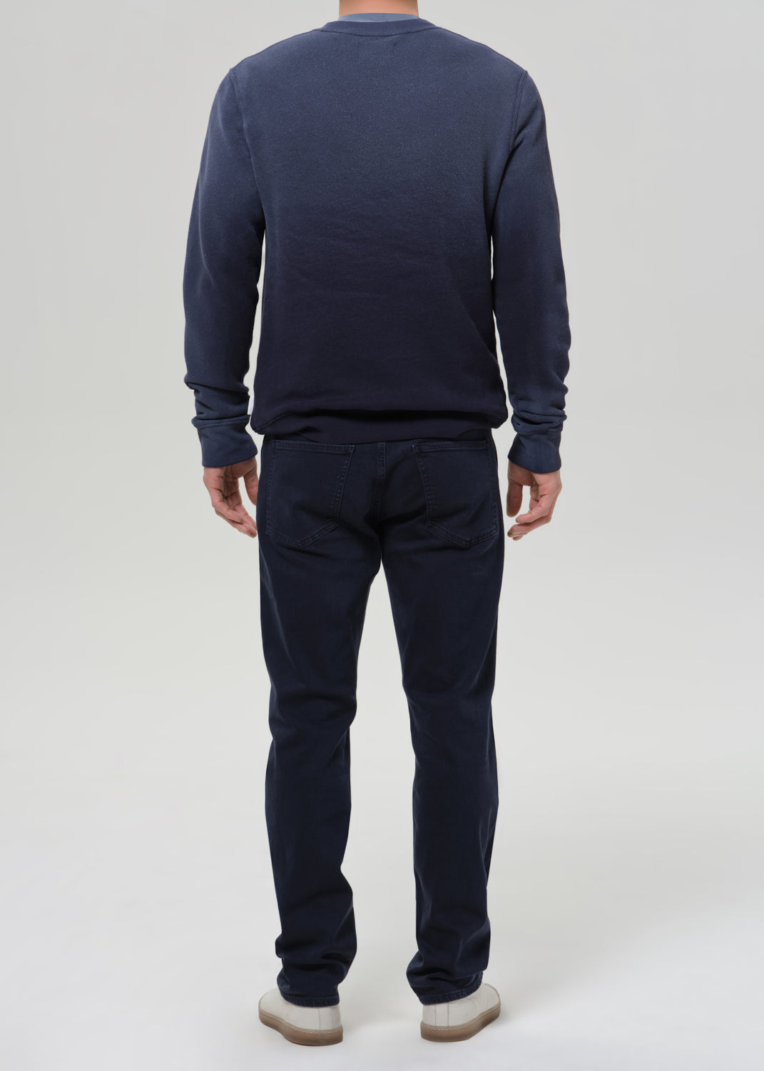 A man viewed from the back wearing a dark navy blue long-sleeve crewneck sweatshirt and matching navy blue pants, paired with white sneakers, against a grey background.