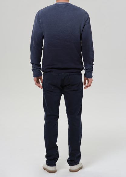 A man viewed from the back wearing a dark navy blue long-sleeve crewneck sweatshirt and matching navy blue pants, paired with white sneakers, against a grey background.