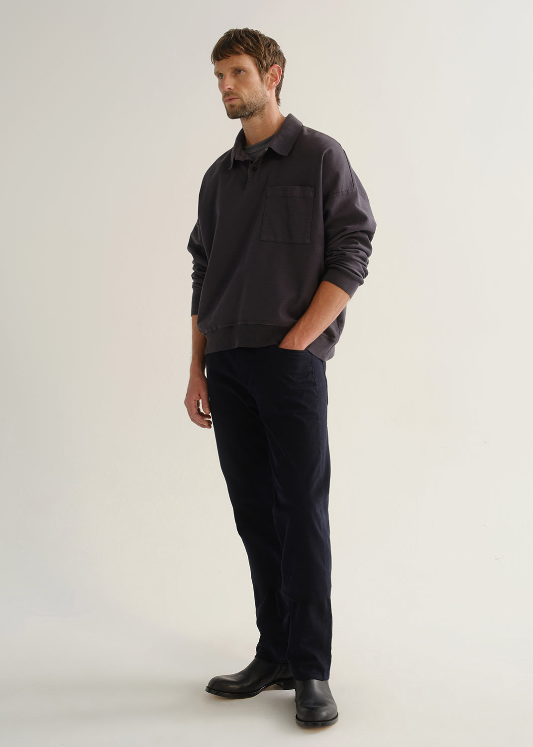 Person wearing dark navy straight leg jeans with a charcoal long sleeve shirt and black boots, angled front full body view on a light grey background.