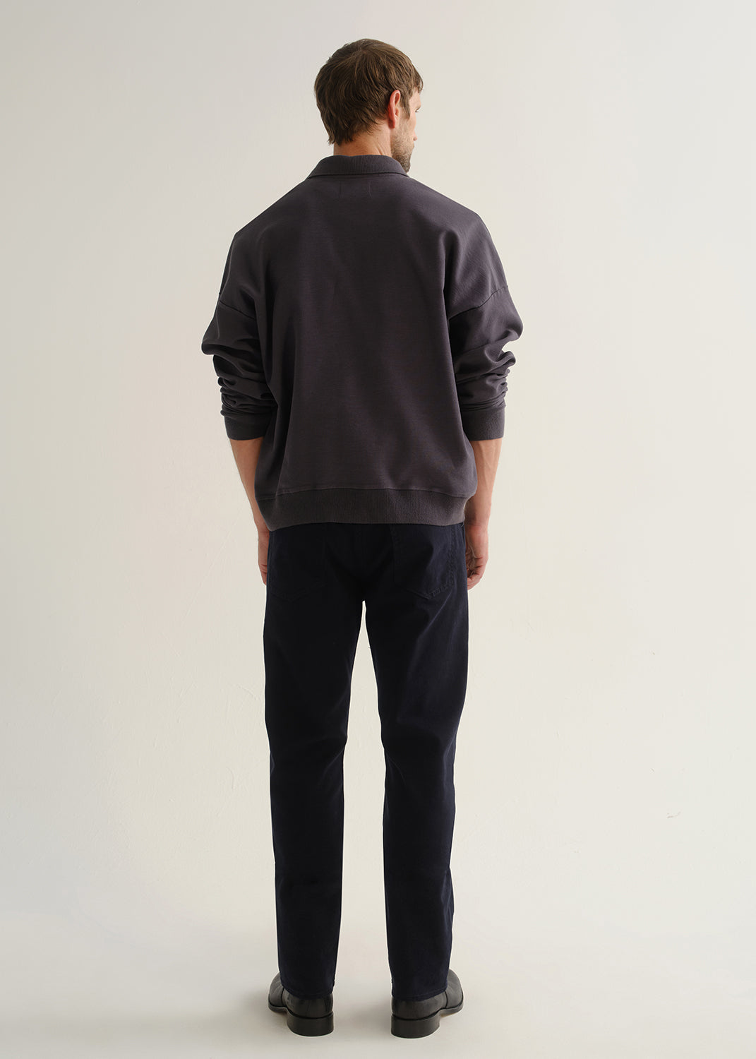 Person wearing dark navy straight leg jeans with a charcoal long sleeve shirt and black boots, back full body view on a light grey background.