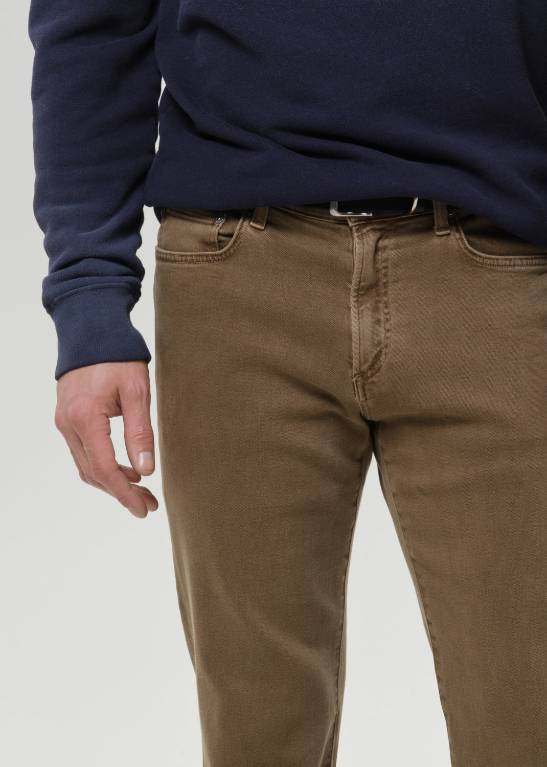 A close-up of a person wearing brown slim-fit jeans and a navy blue sweatshirt, cropped from the chest down against a grey background.