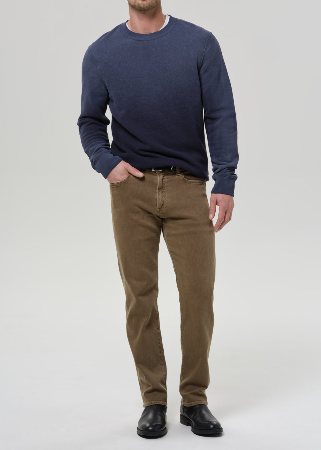A man wearing a navy blue long-sleeve crewneck sweater, brown trousers, and black leather shoes, standing against a grey background.