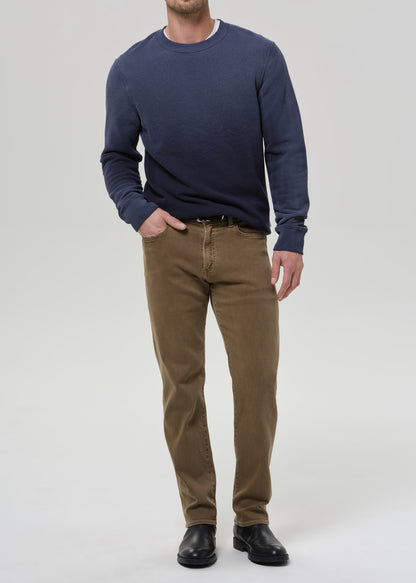 A man wearing a navy blue long-sleeve crewneck sweater, brown trousers, and black leather shoes, standing against a grey background.