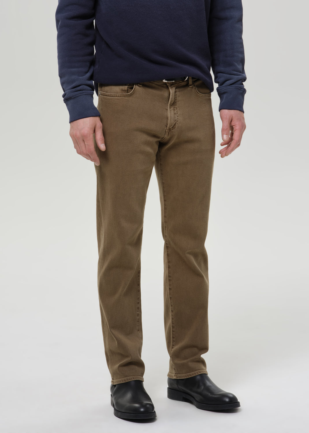 A man wearing slim-fit brown trousers, a navy blue long-sleeve shirt, and black leather boots, standing against a grey background.