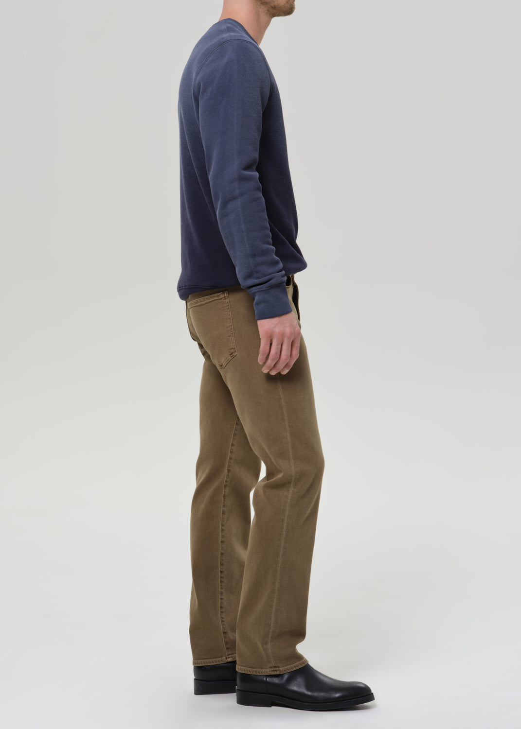 A man shown in profile wearing a navy blue long-sleeve shirt, brown pants, and black shoes, standing against a grey background.