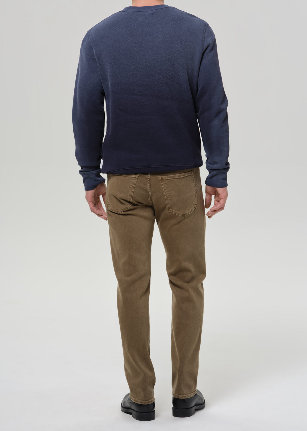 A man viewed from the back wearing a navy blue long-sleeve crewneck sweater and brown trousers, standing against a grey background.