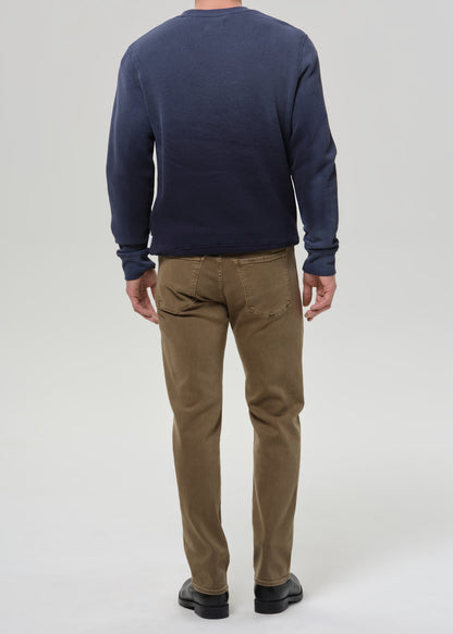 A man viewed from the back wearing a navy blue long-sleeve crewneck sweater and brown trousers, standing against a grey background.