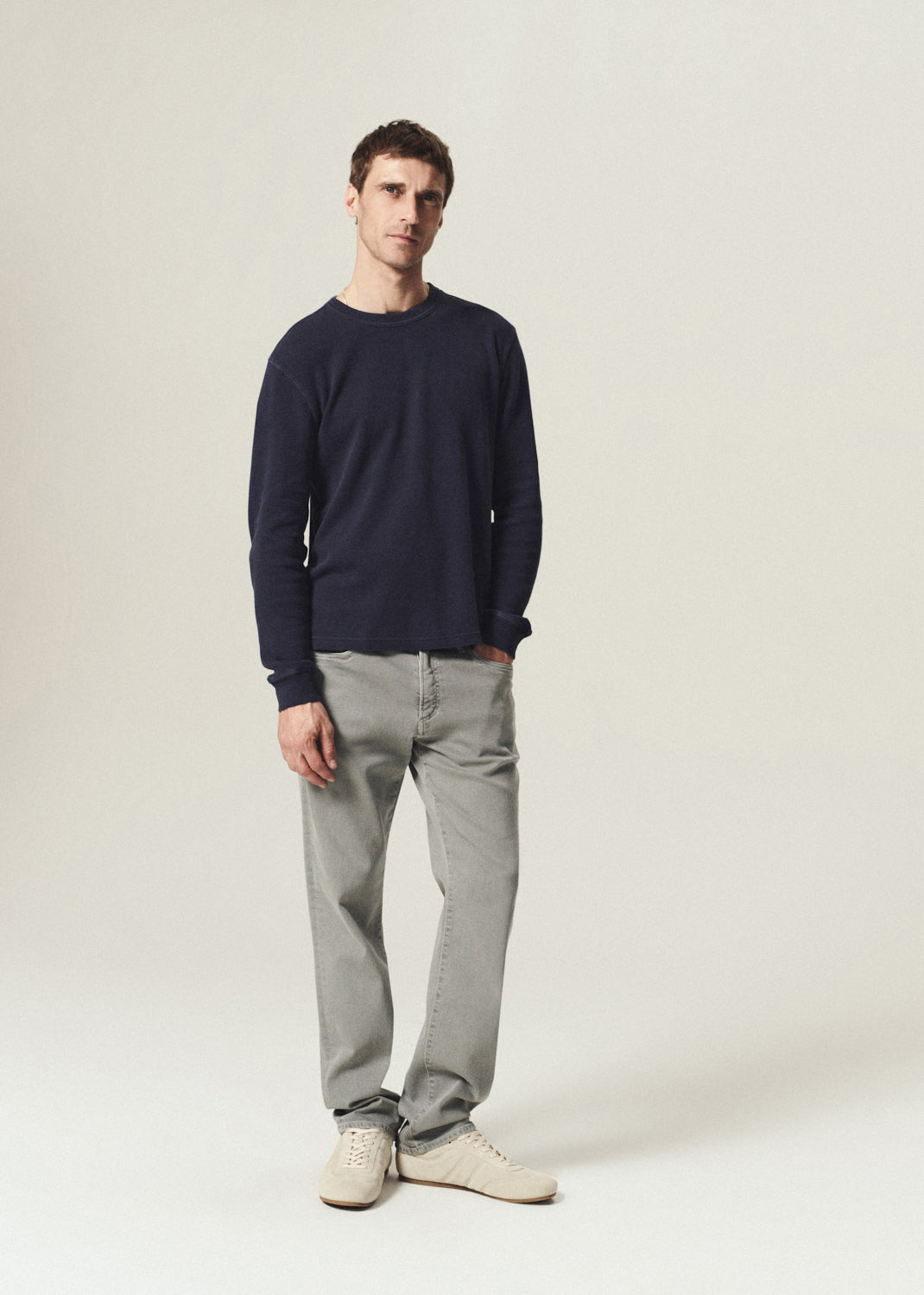 A man modeling a dark navy long-sleeve crewneck sweater, light gray slim-fit jeans, and off-white sneakers against a plain, light beige studio background.