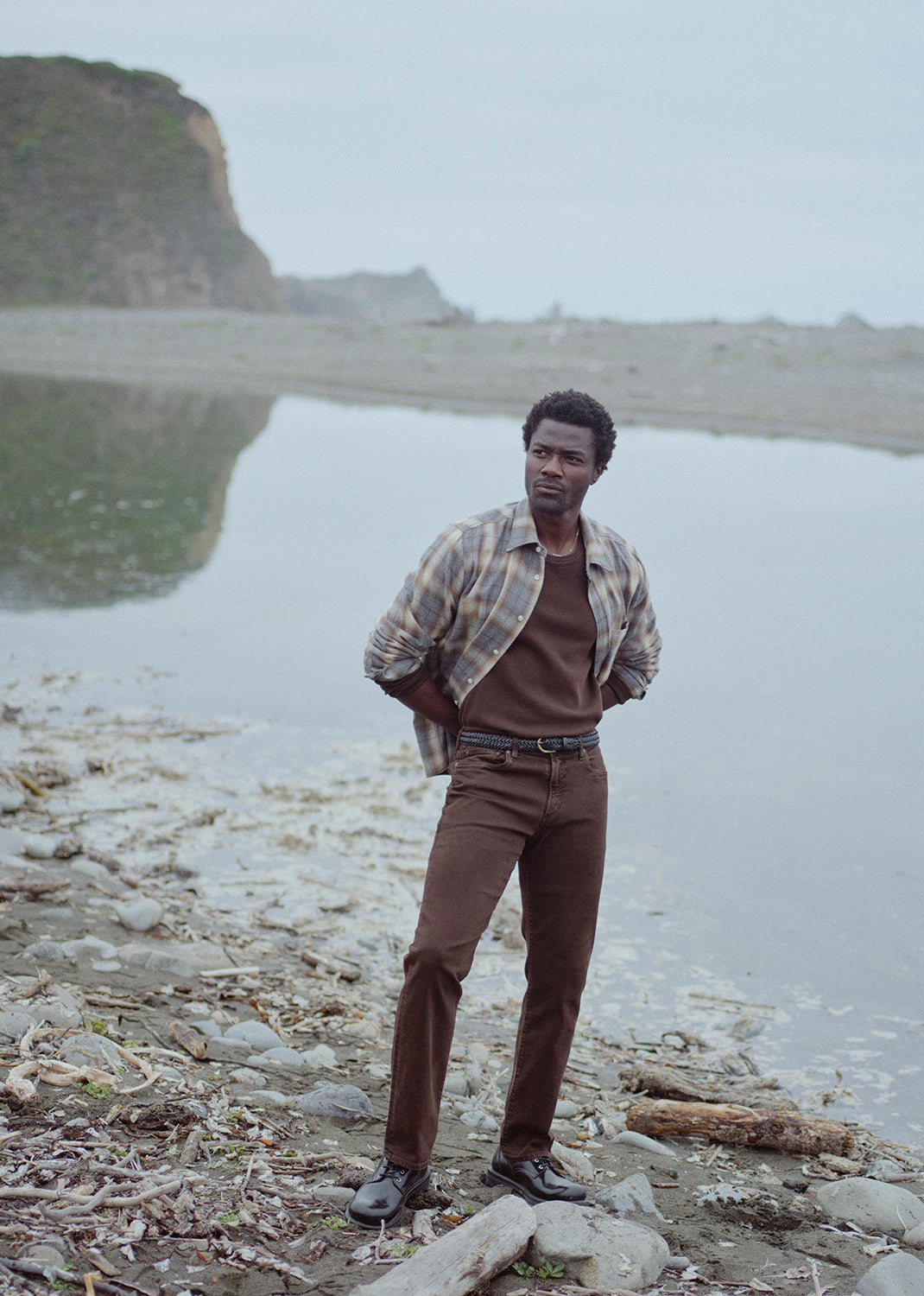 A man stands on a rocky shoreline by calm water, wearing a brown shirt and brown pants with a plaid button-up shirt draped off his shoulders. A large rocky cliff is visible in the background under an overcast sky.