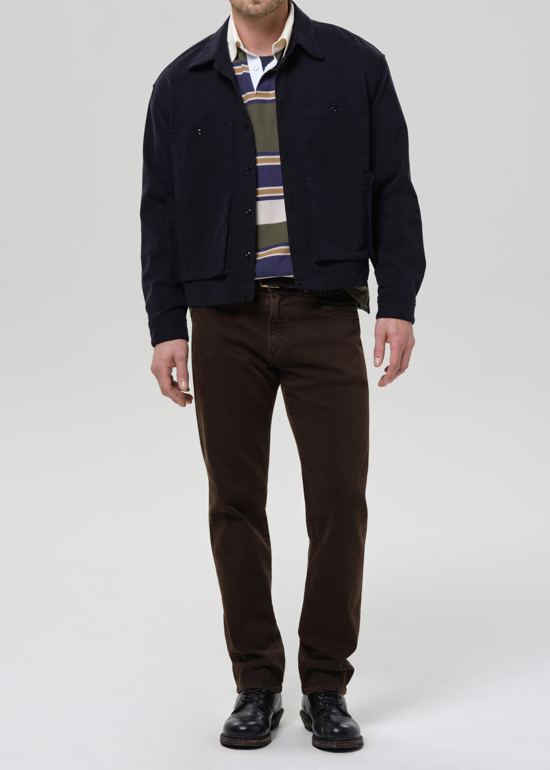 Man wearing a navy jacket, striped shirt, and brown pants on a grey background