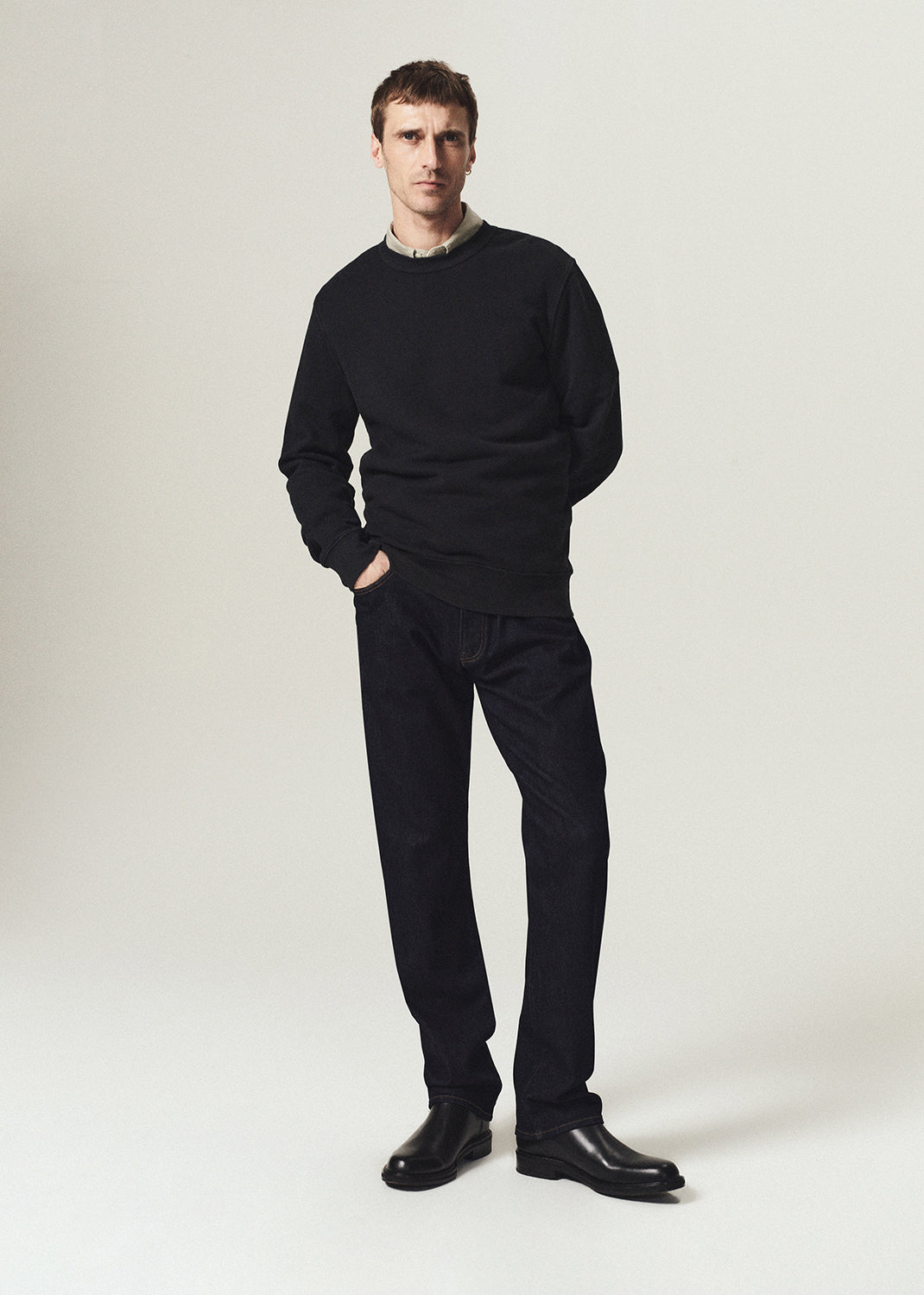 A man modeling a black crewneck sweater, black trousers, and black leather shoes against a light gray studio background.