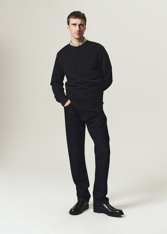 A man modeling a black crewneck sweater, black trousers, and black leather shoes against a light gray studio background.