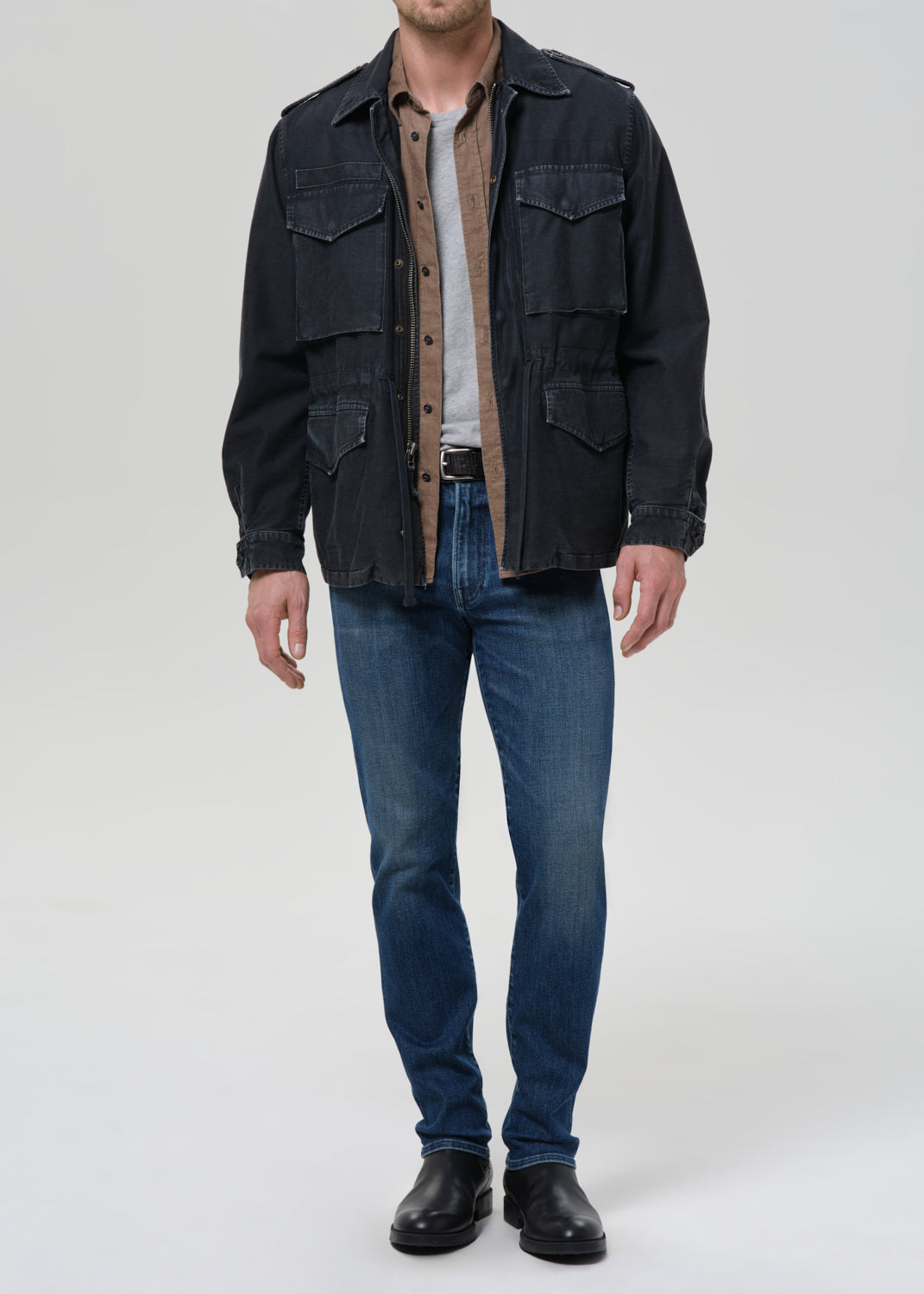 Person wearing a dark denim jacket, brown shirt, and blue jeans on a grey background