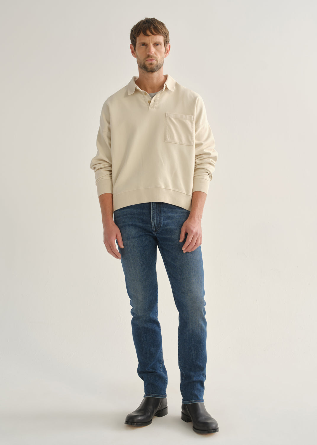 Person wearing medium indigo straight leg jeans with a cream long sleeve collared pullover and black boots, front full body view on a light grey background.