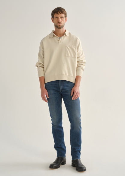 Person wearing medium indigo straight leg jeans with a cream long sleeve collared pullover and black boots, front full body view on a light grey background.