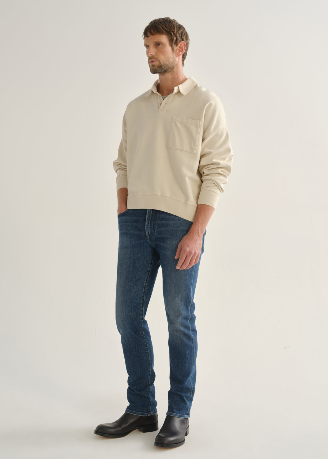 Person wearing medium indigo straight leg jeans with a cream long sleeve collared pullover and black boots, angled front full body view on a light grey background.
