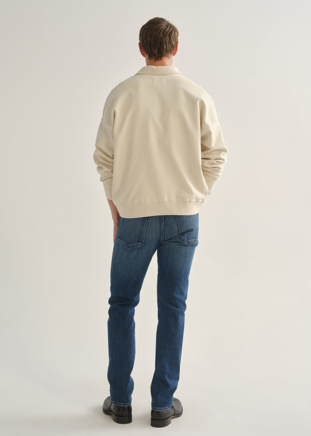 Person wearing medium indigo straight leg jeans with a cream long sleeve collared pullover and black boots, back full body view on a light grey background.