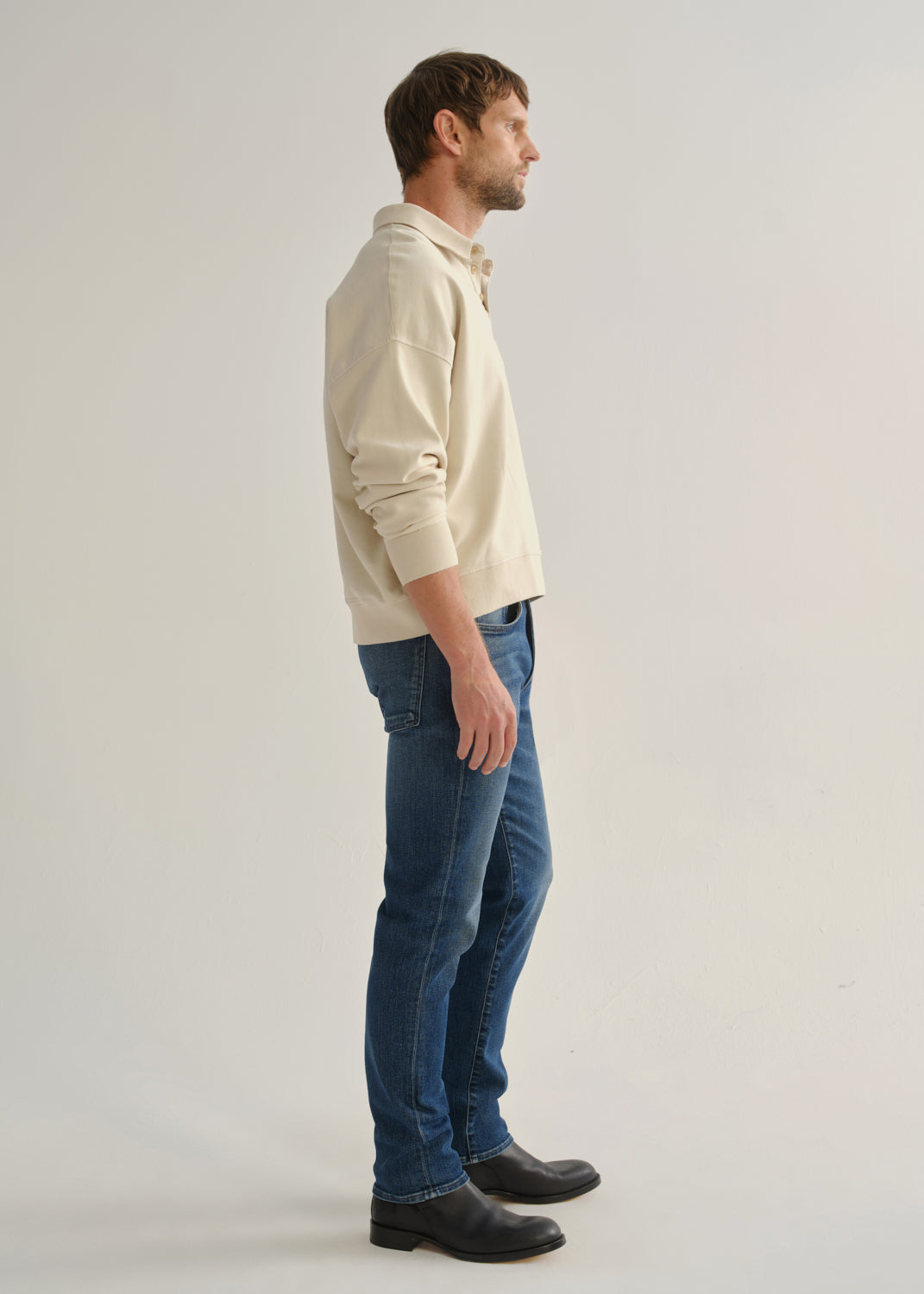 Person wearing medium indigo straight leg jeans with a cream long sleeve collared pullover and black boots, side full body view on a light grey background.