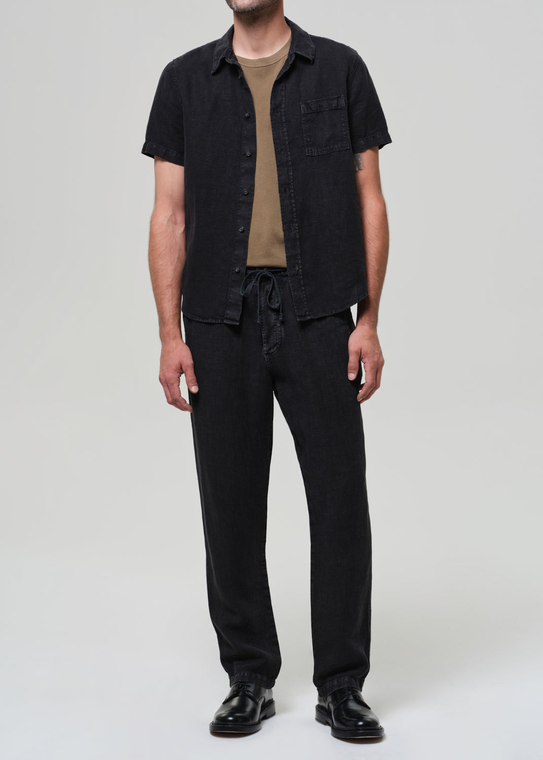Person wearing a black short-sleeve linen shirt over a beige t-shirt, paired with black linen pants on a grey background